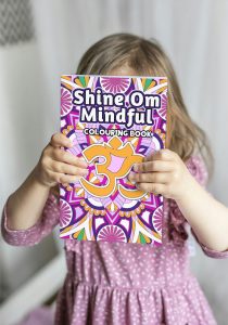 Child holding the Shine Om Mindful Colouring Book upright, showing its bright mandala-style cover in purple, orange, and pink tones — a mindfulness colouring book created by Shine Om to support calm and creativity for kids and families. Learn more about this book crafted for kids and families and purchase a copy at www.shineom.com.au