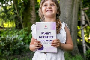 Child standing outdoors in a lush green setting, smiling and holding the Family Gratitude Journal by Shine Om — a mindfulness workbook designed to nurture calm, connection, and kindness through simple daily gratitude rituals. Learn more about the Journal and purchase a copy at www.shineom.com.au